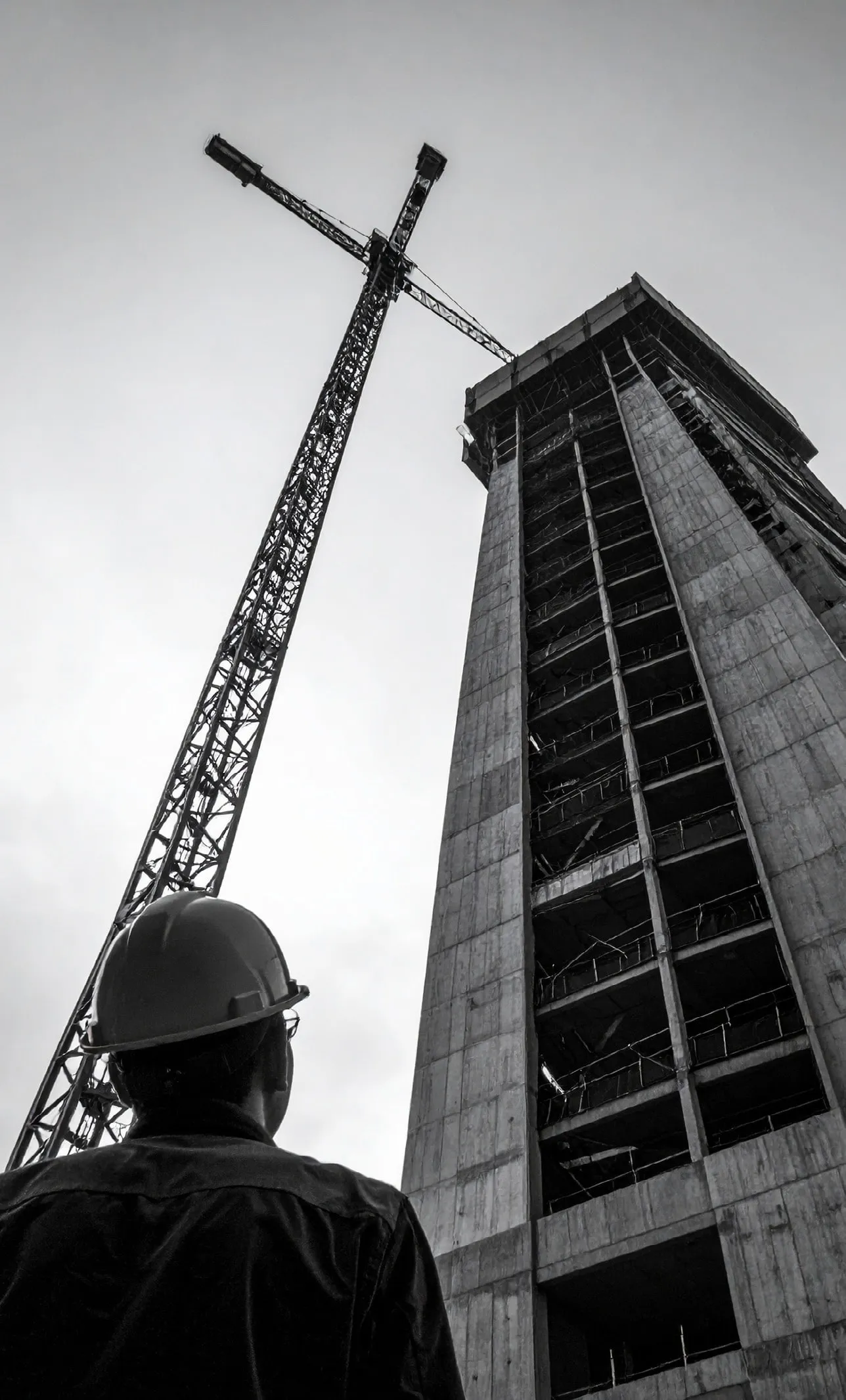 A site manager in a hard hat looks up at a tower under construction and its crane