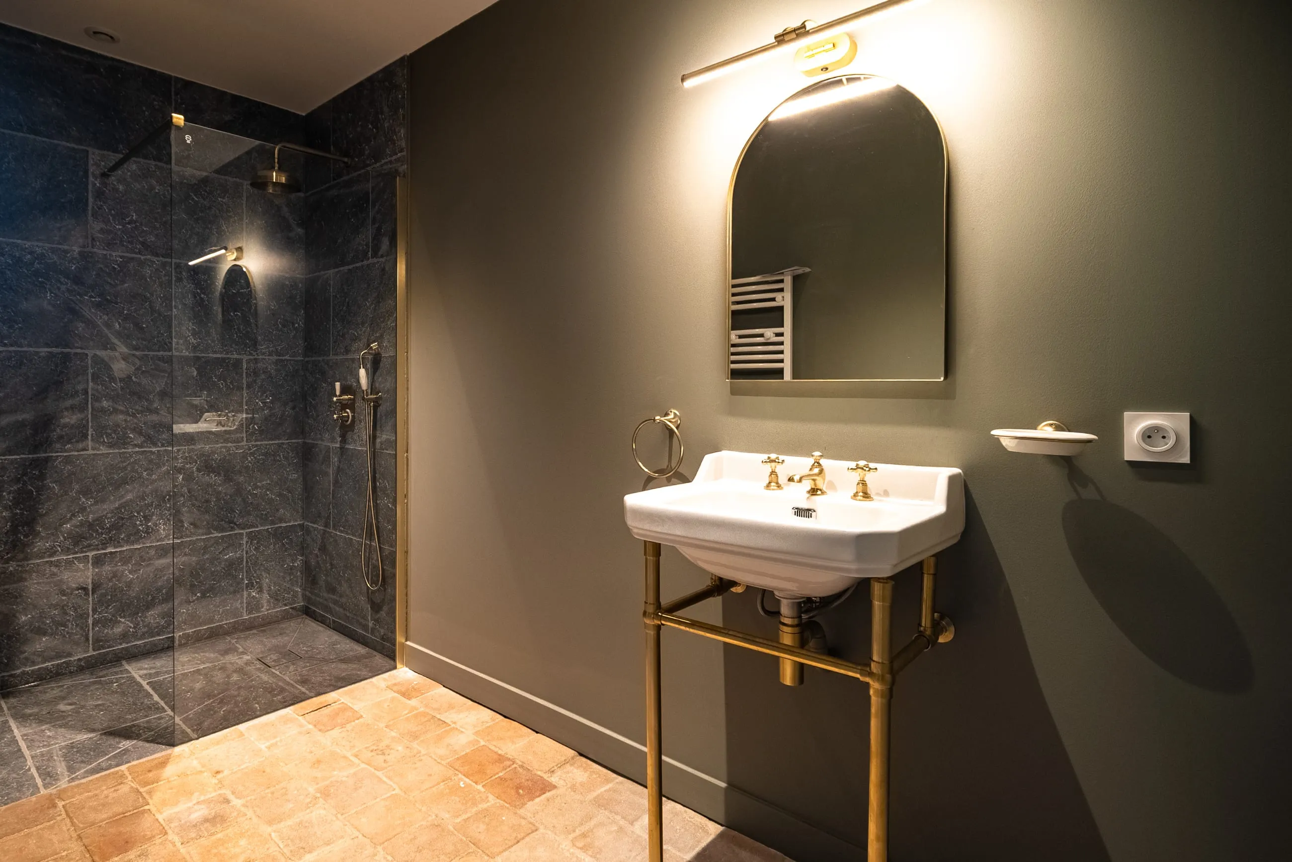 Une salle de bain aux murs vert sourd, vasque sur piètement de laiton et douche en marbre gris