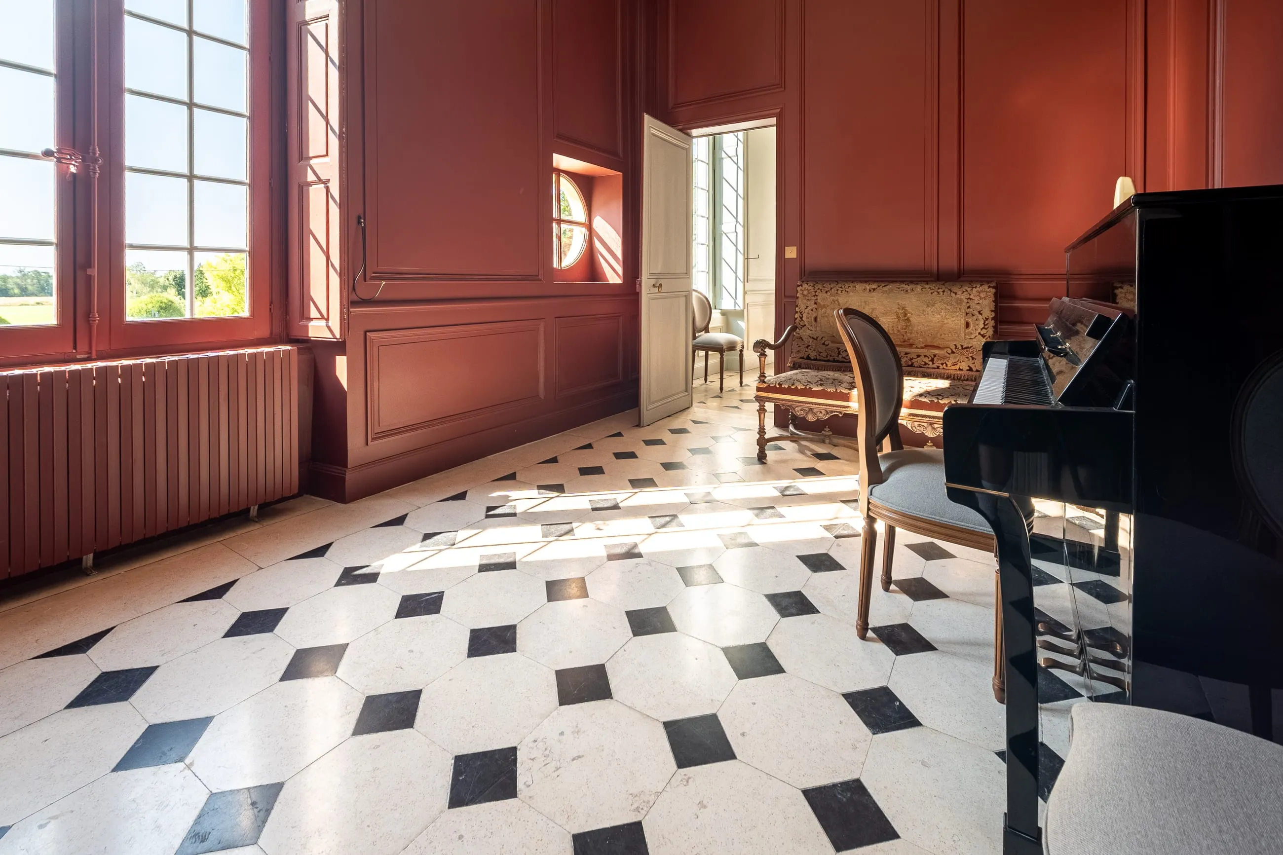 Le grand salon en enfilade, boiseries rouges, sol à cabochons et piano à queue devant les fenêtres